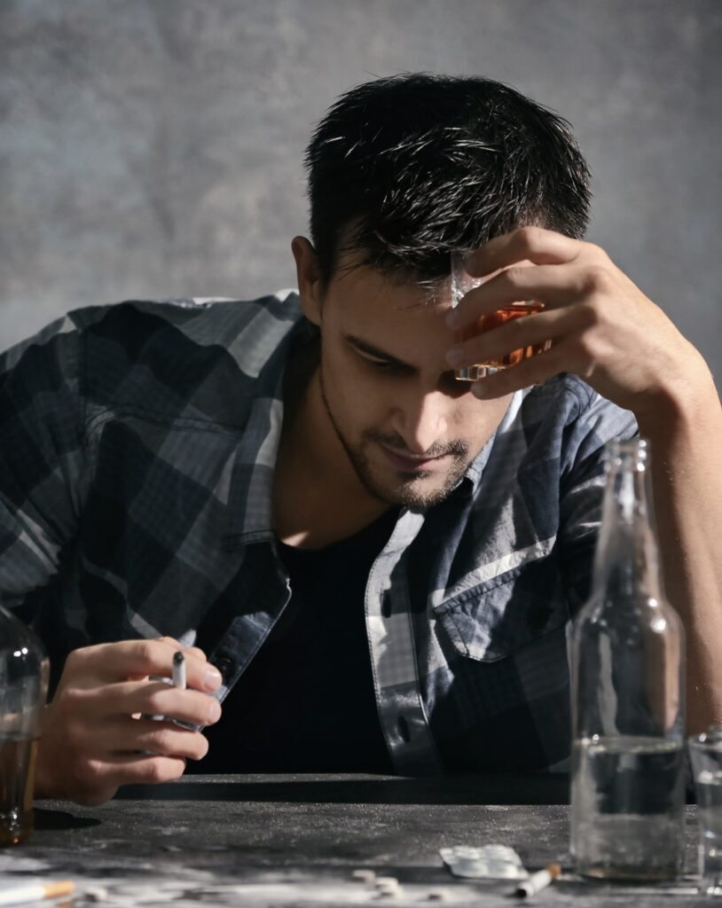 Holding his head in his hands, a man sits at a table with alcohol and pills in front of him