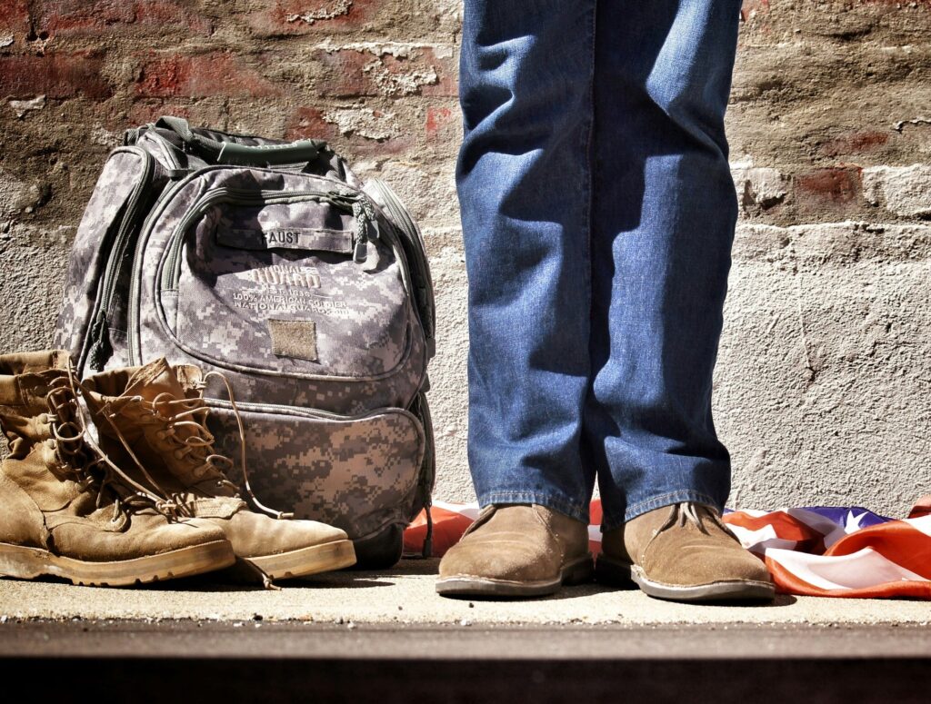 The legs of a person standing next to an army-issue bag, boots, and an American flag, possibly a veteran who could benefit from mental health care.